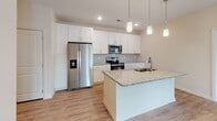 Located on Conductor Circle in Greer, South Carolina, this single-floor home offers a clean, modern aesthetic defined by a bright, neutral palette and consistent light wood-look flooring that flows throughout the main living areas. The open-concept kitchen is a standout feature, showcasing white shaker-style cabinetry extending to the ceiling, granite countertops, a tiled backsplash, and a full suite of stainless steel appliances including a French door refrigerator with ice maker, electric range, and built-in microwave. A large central island with a granite surface and undermount sink serves as both a prep station and casual gathering point, illuminated by stylish pendant lighting above. The spacious living area benefits from vaulted ceilings and glass French doors that invite natural light and provide outdoor access. Bedrooms feature plush carpeting and ceiling fans, with natural light provided by well-positioned windows. The bathrooms are finished with granite-topped white vanities, tile-surround tub-shower combinations, and light wood-look flooring, maintaining the home's cohesive, contemporary style. Additional storage is thoughtfully integrated throughout the layout, contributing to the home's functional and polished overall design.