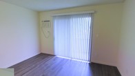 This single-floor space is characterized by its clean, functional layout and practical features. The room showcased in the image is equipped with durable hardwood-style flooring in a rich, dark tone that contrasts neatly with the light yellow-toned walls, lending the space a warm and inviting feel. A large sliding glass door serves as the focal point of the room, allowing an abundance of natural light to filter through the vertical blinds, which provide flexible light control and privacy. The sliding door also offers potential access to an outdoor area. A wall-mounted air conditioning unit is conveniently positioned nearby, ensuring climate comfort throughout the seasons. The neutral wall color and open layout give the space a versatile quality, making it easy to furnish and personalize. The overall ambiance is bright and airy, with the natural light from the sliding door enhancing the sense of openness. The space presents a straightforward, move-in-ready environment suitable for a variety of uses.