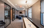Located at 940 Stuyvesant Ave in Union, New Jersey, this contemporary amenity lounge offers a well-appointed, single-floor space designed for both relaxation and social engagement. The interior showcases a refined modern aesthetic, with textured grey wall coverings, tray ceilings with recessed lighting, and plush carpeted flooring throughout.

The space is divided into distinct zones, beginning with a stylish lounge area furnished with sculptural blue accent chairs and a sleek media console topped with decorative accessories, all positioned beneath a wall-mounted flat-screen television. Curated framed artwork adds visual interest to the walls.

A dedicated game area anchors the opposite end of the room, centered around a full-length shuffleboard table beneath a striking multi-arm brass chandelier. Bar-height seating and additional wall art complete the social atmosphere of this zone.

Floor-to-ceiling black-framed glass partitions elegantly define the space while maintaining an open, connected feel. Large windows with roller shades allow natural light to filter in and offer views of the surrounding Union, NJ streetscape. An elevator lobby is visible beyond the glass partitions, suggesting this lounge serves as a premier common area within a larger residential or mixed-use building. The overall ambiance is polished, inviting, and thoughtfully curated for modern community living.