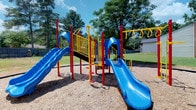 This outdoor playground is a lively, well-equipped recreational space designed to encourage active play for children. The centerpiece is a multi-level play structure built with bold red support poles, bright yellow railings and safety barriers, and vivid blue molded plastic components, creating an eye-catching and energetic aesthetic.

The main structure features two broad blue slides — one larger curved slide and a second straight slide — providing multiple play options. Elevated platforms with yellow guardrails connect the various elements, allowing children to navigate the structure safely. A set of monkey bars extends from a yellow horizontal frame to one side, offering an additional upper-body challenge.

The ground beneath the equipment is covered with a generous layer of wood chip mulch, providing a cushioned, safety-conscious surface throughout the play area. The surrounding landscape includes a well-maintained open lawn and a backdrop of tall, mature deciduous and pine trees that provide natural shade. A building is partially visible at the edge of the property, and fencing encloses the perimeter, contributing to a safe and contained environment ideal for supervised outdoor recreation.