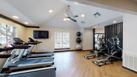 Located at 9818 N MacArthur Blvd in Irving, TX, this fitness center is a thoughtfully designed, single-floor facility offering a comprehensive workout environment. The space features a combination of vaulted and standard ceilings, with recessed lighting and a ceiling fan providing both style and comfort throughout. Herringbone-patterned hardwood flooring runs throughout the space, lending a clean, modern aesthetic that ties the room together.

The cardio zone is well-appointed with multiple treadmills and elliptical machines positioned near large windows, allowing natural light to fill the space and offering views of the surrounding outdoor area. Stability balls are neatly organized nearby, supporting functional fitness routines. The strength training section houses a Life Fitness multi-station cable machine and a Synrgy360 functional training system, alongside a dumbbell rack and adjustable weight bench, catering to a wide range of resistance training needs. A stair climber adds further variety to the equipment selection.

Textured accent walls in warm earth tones provide visual contrast against the light-colored walls, while wall-mounted flat-screen televisions offer entertainment during workouts. Large glass entry doors with black frames bring in additional natural light and connect the interior to an outdoor deck area, creating an open and inviting atmosphere.