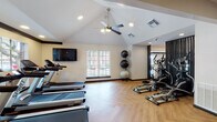 Located at 9818 N MacArthur Blvd in Irving, TX, this fitness center is a thoughtfully designed, single-floor facility offering a comprehensive workout environment. The space features a combination of vaulted and standard ceilings, with recessed lighting and a ceiling fan providing both style and comfort throughout. Herringbone-patterned hardwood flooring runs throughout the space, lending a clean, modern aesthetic that ties the room together.

The cardio zone is well-appointed with multiple treadmills and elliptical machines positioned near large windows, allowing natural light to fill the space and offering views of the surrounding outdoor area. Stability balls are neatly organized nearby, supporting functional fitness routines. The strength training section houses a Life Fitness multi-station cable machine and a Synrgy360 functional training system, alongside a dumbbell rack and adjustable weight bench, catering to a wide range of resistance training needs. A stair climber adds further variety to the equipment selection.

Textured accent walls in warm earth tones provide visual contrast against the light-colored walls, while wall-mounted flat-screen televisions offer entertainment during workouts. Large glass entry doors with black frames bring in additional natural light and connect the interior to an outdoor deck area, creating an open and inviting atmosphere.
