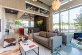 Located in Atlanta, GA, this single-floor space combines contemporary design with artistic flair to create a dynamic, multi-functional environment. The layout flows across polished concrete floors, connecting distinct zones that each offer their own character and purpose.

The main lounge area is defined by expansive floor-to-ceiling glass walls that flood the interior with natural light while framing direct views of the surrounding urban streetscape. Comfortable, modern gray sofas and armchairs are arranged atop a colorful area rug, creating an inviting gathering space beneath soaring ceilings. A sculptural pendant light adds a warm focal point overhead.

An adjacent seating area features a raised platform with light hardwood flooring, anchored by a wall-mounted display screen and dark wood paneling. The ceiling above is adorned with a geometric hexagonal tile pattern in red, blue, and gray tones, adding a playful architectural detail.

A bold, large-scale floral mural by artist Peter Ferber commands one wall, accompanied by curated artwork and a built-in bookshelf below, infusing the space with vibrant creative energy. Cowhide ottomans and eclectic furnishings further enhance the artistic atmosphere.

The kitchen area features sleek, full stainless steel cabinetry and appliances, white countertops, a subway tile backsplash, and a wall-mounted television, combining functionality with a professional, modern aesthetic.