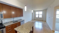 This single-floor unit showcases a thoughtfully designed open-concept layout that seamlessly connects the kitchen and living areas. The kitchen is equipped with rich, warm-toned wood cabinetry paired with sleek brushed-metal hardware, providing ample storage. A generous granite-topped island anchors the space, featuring distinctive speckled patterning and offering both additional workspace and casual seating. The countertops continue the granite theme, complemented by a classic white subway tile backsplash and a stainless steel sink. A stainless steel dishwasher is integrated neatly beneath the counter. Two elongated cylindrical pendant lights hang above the island, casting focused, warm illumination over the workspace. Light hardwood flooring runs throughout, contributing to a cohesive and airy feel across the space. The living area benefits from a large, multi-pane window that allows abundant natural light to fill the room, brightening the clean white walls. The elevated ceiling height enhances the overall sense of openness, creating a spacious and inviting atmosphere throughout this well-appointed unit.