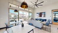 This thoughtfully designed single-floor community clubhouse offers a welcoming and functional gathering space with a contemporary aesthetic throughout. Soaring ceilings amplify the sense of openness, while expansive floor-to-ceiling sliding glass doors and clerestory windows flood the interior with natural light and frame views of the outdoor patio and surrounding neighborhood.

The main lounge area is anchored by a comfortable seating arrangement — featuring neutral-toned sofas and accent chairs — centered around a warm wooden coffee table, with a wall-mounted flat-screen TV and a sleek navy credenza completing the media wall. An oversized ceiling fan with broad-sweep blades adds both function and visual drama overhead, complemented by bold black-and-gold pendant lighting.

The kitchenette is well-appointed with dark wood cabinetry, quartz countertops, a deep undermount sink with a brushed gold faucet, open wood shelving, and stainless steel appliances including a full-size refrigerator. The adjacent dining area accommodates multiple tables with modern marble-top surfaces, sleek black chairs, and striking pendant fixtures overhead.

Decorative wall panels with geometric relief texture add an artistic accent near the entrance, where large glass-panel doors open directly to the exterior. Large-format light tile flooring and crisp white walls tie the entire space together in a clean, cohesive design.