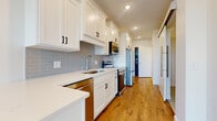 This single-floor space is designed with a clean, contemporary aesthetic that balances functionality and style. The galley-style kitchen is a standout feature, outfitted with crisp white shaker cabinetry accented by matte black hardware, light quartz countertops, and a grey subway tile backsplash. Stainless steel appliances — including a range, microwave, dishwasher, and refrigerator — provide a polished, modern finish. Recessed lighting illuminates the kitchen corridor, which flows seamlessly toward the living areas.

Warm, wood-tone luxury vinyl plank flooring extends throughout the entire unit, lending continuity and a welcoming feel to every room. The living area is bright and open, with white walls and clean lines creating a versatile, neutral canvas. A wall-mounted HVAC unit provides climate control, and a ceiling fan is present in the living space.

Natural light is a defining feature, with a glass-paneled exterior door opening to a private balcony, complemented by a large adjacent window — together flooding the living area with daylight and offering outdoor access. A sliding barn-style door adds a modern architectural detail to the interior layout. The overall ambiance is fresh, minimal, and move-in ready.