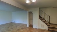 This two-story space is characterized by its warm parquet flooring on the main level, which adds a classic, textured feel to the interior. The main living area is open and versatile, with neutral white walls that create a bright, clean backdrop. A built-in closet with open shelving is conveniently tucked into the wall, offering practical storage directly off the main living space. Overhead, a multi-bulb ceiling light fixture provides functional illumination. A staircase with a sleek metal railing leads to the second floor, adding a structural focal point to the room. A floor-level vent indicates a central HVAC system serving the space. The layout is straightforward and efficient, lending itself well to flexible furniture arrangements. The overall ambiance is understated and functional, with the two-level design providing a natural separation between living areas across floors.