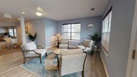 Located at 1830 Magwood Rd in Charleston, SC, this single-story community clubhouse offers a thoughtfully designed gathering space with multiple distinct areas for socializing and recreation. The interior is finished with soft blue-gray walls, a combination of hardwood-style flooring and patterned carpet tiles, and recessed lighting throughout, creating a welcoming and cohesive atmosphere.

The main lounge features two large sofas, accent chairs, and a central coffee table, all arranged around a wall-mounted flat-screen TV and a decorative sideboard. Adjacent seating areas provide quieter spots with armchairs grouped around small tables, accented by potted tropical plants and decorative mirrors. Large windows dressed with wood-tone blinds allow ample natural light to fill each area.

A dedicated game room houses a full-size pool table alongside additional seating, with direct access to an outdoor area via French doors. The mail center is a functional and spacious room featuring an extensive bank of individual resident mailboxes along one wall, complemented by a small café-style table and chairs. The layout flows openly between zones, with columns providing architectural definition. The overall design balances practicality with comfort, making this clubhouse a versatile hub for community life.
