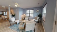 Located at 1830 Magwood Rd in Charleston, SC, this single-story community clubhouse offers a thoughtfully designed gathering space with multiple distinct areas for socializing and recreation. The interior is finished with soft blue-gray walls, a combination of hardwood-style flooring and patterned carpet tiles, and recessed lighting throughout, creating a welcoming and cohesive atmosphere.

The main lounge features two large sofas, accent chairs, and a central coffee table, all arranged around a wall-mounted flat-screen TV and a decorative sideboard. Adjacent seating areas provide quieter spots with armchairs grouped around small tables, accented by potted tropical plants and decorative mirrors. Large windows dressed with wood-tone blinds allow ample natural light to fill each area.

A dedicated game room houses a full-size pool table alongside additional seating, with direct access to an outdoor area via French doors. The mail center is a functional and spacious room featuring an extensive bank of individual resident mailboxes along one wall, complemented by a small café-style table and chairs. The layout flows openly between zones, with columns providing architectural definition. The overall design balances practicality with comfort, making this clubhouse a versatile hub for community life.