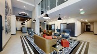 This contemporary lobby lounge makes an immediate impression with its soaring double-height ceilings and open, airy layout. Large mezzanine-level windows overlook the main floor, adding to the sense of verticality and visual depth throughout the space. Pendant lighting in sleek black cone shades descends from the ceiling, casting focused illumination across the thoughtfully curated seating arrangements below. The lounge area is anchored by a bold black-and-white patterned area rug, over which a mix of tan leather sofas, coral accent chairs, and striking blue wingback chairs are artfully arranged. Red cube ottomans and a gold-framed coffee table add vibrant pops of color and a touch of glam. Exposed concrete columns punctuate the space, lending an industrial edge that contrasts beautifully with the refined furnishings. Elevator banks are conveniently positioned at the rear, framed by dark doors that complement the overall neutral palette. A bar or reception counter area is visible to the left, suggesting additional amenity space. The overall ambiance is polished and modern, balancing functionality with a strong design aesthetic that sets a welcoming tone for the building.