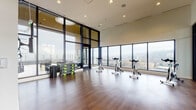 This elevated fitness studio is designed to support a wide range of workout activities within a bright, open, and modern environment. The space features rich hardwood-style flooring throughout, lending warmth to an otherwise sleek, contemporary aesthetic. Soaring ceilings and expansive floor-to-ceiling windows wrap around the room, flooding the interior with natural light and offering sweeping panoramic views of the surrounding cityscape and rolling hills beyond.

The main open area is generously proportioned, providing ample room for yoga, stretching, or group fitness classes. Several spinning bikes are arranged near the window-facing wall, allowing users to exercise while taking in the elevated city views. A well-stocked equipment rack along one wall holds an assortment of kettlebells, dumbbells, resistance tools, a stability ball, and exercise mats, catering to diverse training needs. A wall-mounted flat-screen display provides added functionality for guided workouts or instructional content.

Adjoining the main studio via sleek black-framed glass partition walls is a separate cardio and strength training area, visible from within the studio and equipped with treadmills, cable machines, and additional cardio equipment. Pendant lighting and recessed ceiling fixtures ensure consistent illumination throughout both zones, creating a cohesive and well-appointed fitness environment.