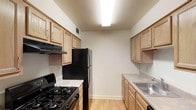 This single-floor space showcases a galley-style kitchen designed for efficient use of space. The kitchen features light oak-toned cabinetry lining both walls, providing ample storage through a combination of upper and lower cabinets. Black appliances, including a gas range and a full-sized refrigerator, create a contrast against the lighter cabinetry and neutral wall tones. A stainless steel sink is positioned along the right-hand wall, accompanied by a countertop offering functional prep space. Light wood laminate flooring runs the length of the kitchen, adding warmth to the area. Overhead, a flush-mount fluorescent light fixture provides consistent, practical illumination throughout the space. A ventilation hood above the range ensures proper airflow while cooking. The layout allows for straightforward movement between the cooking, prep, and cleaning zones, making it a practical and well-organized kitchen. The neutral color palette of beige walls and light wood tones throughout creates a clean, understated aesthetic that is easy to personalize.