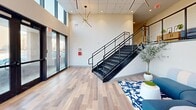 Located at 18545 Hilliard Blvd in Rocky River, Ohio, this contemporary commercial lobby delivers a striking first impression through its modern architectural design and thoughtful interior details. The entrance area is defined by dramatic floor-to-ceiling glass curtain walls framed in sleek dark metal, flooding the space with abundant natural light and offering open views of the surrounding landscape. Light-toned wood-look flooring runs throughout the ground level, complementing the clean, neutral color palette of the walls and ceiling.

A sculptural open-riser staircase with a bold black steel frame and horizontal cable railings serves as a central architectural feature, connecting the main floor to an upper mezzanine level. The mezzanine overlooks the lobby below and is outfitted with carpeted flooring, built-in cabinetry in a warm wood finish, and comfortable seating, creating an intimate secondary lounge or waiting area.

On the main level, the lounge area is furnished with a curved modular sectional sofa in blue and white tones, anchored by a patterned area rug. Eye-catching starburst pendant light fixtures in brushed gold and matte black are suspended from the ceiling, adding a refined, modern aesthetic. Potted greenery throughout softens the contemporary design elements and enhances the welcoming ambiance of this well-appointed commercial space.