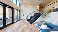 Located at 18545 Hilliard Blvd in Rocky River, Ohio, this contemporary commercial lobby delivers a striking first impression through its modern architectural design and thoughtful interior details. The entrance area is defined by dramatic floor-to-ceiling glass curtain walls framed in sleek dark metal, flooding the space with abundant natural light and offering open views of the surrounding landscape. Light-toned wood-look flooring runs throughout the ground level, complementing the clean, neutral color palette of the walls and ceiling.

A sculptural open-riser staircase with a bold black steel frame and horizontal cable railings serves as a central architectural feature, connecting the main floor to an upper mezzanine level. The mezzanine overlooks the lobby below and is outfitted with carpeted flooring, built-in cabinetry in a warm wood finish, and comfortable seating, creating an intimate secondary lounge or waiting area.

On the main level, the lounge area is furnished with a curved modular sectional sofa in blue and white tones, anchored by a patterned area rug. Eye-catching starburst pendant light fixtures in brushed gold and matte black are suspended from the ceiling, adding a refined, modern aesthetic. Potted greenery throughout softens the contemporary design elements and enhances the welcoming ambiance of this well-appointed commercial space.