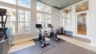 Located at 104 Herrings Green Loop in Mooresville, NC, this fitness center offers a generously sized, single-floor layout designed to support a full range of workout routines. The space is defined by its striking two-toned design — crisp white walls contrast with a bold dark teal ceiling accented by recessed lighting, creating a modern and energizing atmosphere throughout.

The main cardio zone is bathed in natural light from a dramatic wall of tall, multi-pane windows that overlook the surrounding community, giving the space an open, airy feel. Multiple treadmills and elliptical machines are positioned to take full advantage of these views, alongside wall-mounted flat-screen TVs for entertainment during workouts.

A dedicated strength training area features a cable and functional trainer system, a multi-station weight rack, adjustable benches, and a comprehensive selection of dumbbells displayed along a full-length wall mirror that amplifies the sense of space. Pull-up bars and additional cable attachments provide versatility for upper-body training.

A separate cycling alcove houses multiple spin bikes equipped with digital screens, set on rubberized floor mats and framed by additional windows allowing natural light to flow in. The flooring throughout transitions between rubber sport tiles and carpet, offering appropriate surfaces for each activity zone. Together, these elements make this fitness center a well-appointed and functional amenity.