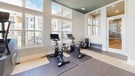 Located at 104 Herrings Green Loop in Mooresville, NC, this fitness center offers a generously sized, single-floor layout designed to support a full range of workout routines. The space is defined by its striking two-toned design — crisp white walls contrast with a bold dark teal ceiling accented by recessed lighting, creating a modern and energizing atmosphere throughout.

The main cardio zone is bathed in natural light from a dramatic wall of tall, multi-pane windows that overlook the surrounding community, giving the space an open, airy feel. Multiple treadmills and elliptical machines are positioned to take full advantage of these views, alongside wall-mounted flat-screen TVs for entertainment during workouts.

A dedicated strength training area features a cable and functional trainer system, a multi-station weight rack, adjustable benches, and a comprehensive selection of dumbbells displayed along a full-length wall mirror that amplifies the sense of space. Pull-up bars and additional cable attachments provide versatility for upper-body training.

A separate cycling alcove houses multiple spin bikes equipped with digital screens, set on rubberized floor mats and framed by additional windows allowing natural light to flow in. The flooring throughout transitions between rubber sport tiles and carpet, offering appropriate surfaces for each activity zone. Together, these elements make this fitness center a well-appointed and functional amenity.