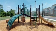 This outdoor playground offers a well-equipped recreational space designed to engage children of various ages and activity levels. The centerpiece is a multi-level play structure finished in dark brown and green tones, blending naturally with the outdoor setting. The structure features multiple play elements, including a curved climbing wall with tactile handholds, a spiral slide, a straight slide, a bridge walkway with safety railings, and ladder-style step accesses. Interactive panels, including a colorful activity board, are integrated into the structure to encourage imaginative and sensory play. The entire play area is surfaced with a generous layer of wood chip mulch, providing a cushioned, safety-conscious ground cover beneath and around all equipment. The playground is set within a spacious open lawn area under a clear blue sky, allowing ample room for free play beyond the main structure. A classic-style lamppost stands nearby, suggesting the area is accessible during evening hours as well. Residential buildings are visible in the background, indicating this playground serves as a community amenity. The overall environment is bright, clean, and inviting, offering a safe and engaging outdoor destination for children and families.