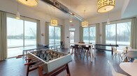 Located at 720 Michigan Ave in Columbus, Ohio, this single-floor community amenity space is designed for socializing and recreation. The game room is a standout feature, boasting soaring ceilings with exposed industrial ductwork, stylish geometric pendant lighting, and rich dark hardwood flooring throughout. Expansive floor-to-ceiling windows dressed with elegant linen drapes flood the room with natural light and offer open views of the surrounding neighborhood. The space is well-equipped with a foosball table, a ping pong table, a high-top bar table with stools, and a small bistro table with modern acrylic chairs — providing ample options for both competitive play and casual gathering. Adjacent to the game area is a vibrant lounge seating area featuring a bold teal channeled banquette, modern round tables, and multiple wall-mounted flat-screen TVs, creating an inviting space for relaxed viewing and conversation. A fully equipped kitchenette rounds out the amenities, outfitted with dark cabinetry, white countertops, a stainless steel refrigerator, microwave, sink, and dishwasher — ideal for hosting events or casual get-togethers. The overall ambiance is modern, bright, and thoughtfully designed to encourage community engagement.