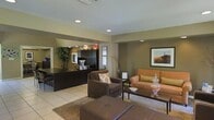 This single-story space functions as a combined reception and lounge area, thoughtfully designed to accommodate both administrative tasks and guest comfort. The reception desk, finished in dark wood cabinetry, anchors one side of the room and provides a organized workspace with built-in shelving and computer workstations. Recessed lighting throughout the ceiling illuminates the space evenly, complementing the neutral, earthy wall tones in warm olive and taupe hues.

The seating area is furnished with a rich brown upholstered armchair accented by a patterned throw pillow, alongside a tan sofa with coordinating cushions, arranged around a dark wood coffee table. Decorative elements such as a glass apothecary jar, a shallow bowl, and a tall floor vase with dried branches add refined touches throughout. Framed artwork featuring desert landscape photography adorns the walls, reinforcing a Southwestern aesthetic.

Light-toned ceramic tile flooring extends across the space, partially covered by a neutral area rug beneath the lounge furniture. A potted tree near the reception desk introduces a natural element. The overall ambiance is professional yet approachable, balancing functionality with comfortable hospitality.