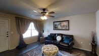 Located in Lubbock, Texas, this single-story home offers a welcoming and well-appointed interior designed for comfortable everyday living. The living area serves as the heart of the home, showcasing tile flooring laid in a neutral tone that provides both durability and easy maintenance. A ceiling fan with a built-in light fixture is centrally mounted, offering efficient air circulation and ambient lighting throughout the space. Natural light filters in through a window dressed with layered curtains — a combination of sheer panels and patterned drapes — adding warmth and privacy to the room. The living area is thoughtfully arranged, featuring a dark leather sofa accented with decorative teal and cream throw pillows, complemented by a tufted, floral-patterned ottoman that serves as a stylish centerpiece. A classic landscape painting in a dark ornate frame adorns the wall, contributing to the room's traditional aesthetic. A decorative area rug anchors the seating arrangement, adding texture and visual interest to the space. The overall ambiance of this home is warm, intimate, and classic, making it a comfortable retreat in the heart of Lubbock.