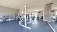 This fitness center is a generously sized, single-floor exercise facility designed to accommodate a full range of workout routines. The space is entered through a tiled entry corridor equipped with a water fountain and restroom access, transitioning into the main workout area with durable, dark rubber flooring suited for heavy equipment use.

The exercise room is bathed in natural light through large, multi-pane windows along the walls, offering outdoor views and an open, airy ambiance. Neutral gray walls and recessed lighting contribute to a clean, focused atmosphere throughout. A large wall mirror expands the sense of space and supports proper form during strength training.

The strength training area is well-appointed with a multi-station cable machine, a squat/power rack, bench press setup, a leg press machine, adjustable benches, and a dumbbell rack stocked with a full range of free weights. The cardio section features multiple elliptical trainers, treadmills, and a stationary bike, all positioned to take advantage of the natural light and outdoor views. A wall-mounted television provides entertainment for cardio sessions.

The vaulted ceiling in portions of the room adds to the expansive feel, making this fitness center a comprehensive and well-organized space for both cardiovascular and resistance training.