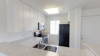 This single-floor space presents a clean, modern aesthetic anchored by a thoughtfully designed kitchen. The kitchen features crisp white cabinetry that extends to the ceiling, maximizing storage capacity, and is paired with light granite-style countertops that lend a polished, contemporary feel. A stainless steel double-basin sink sits prominently at the counter, complemented by a full suite of appliances including a black refrigerator, a stainless steel electric range with oven, and an over-the-range microwave. The layout follows an efficient galley-style configuration, optimizing workflow and usability. A window positioned at the end of the kitchen invites natural light into the space, brightening the white surfaces and contributing to an open, airy atmosphere. Recessed flush ceiling lighting provides consistent illumination throughout. The overall color palette of white walls, white cabinetry, and light countertops creates a cohesive, neutral environment that feels clean and inviting. The space reflects a functional design philosophy, offering practical features in a well-organized, single-level layout suited for everyday living.