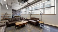 Located at 22 S Saint Clair St in the heart of downtown Dayton, Ohio, this single-floor commercial space blends industrial character with functional, collaborative design. The open layout is anchored by exposed ceiling ductwork, suspended linear LED lighting, and structural concrete columns that reinforce the space's loft-like aesthetic. Patterned modular carpet tiles in muted earth and slate tones ground the interior throughout.

The space is divided into distinct functional zones. A lounge area features rich brown leather sofas and loveseats arranged around a reclaimed wood-and-iron coffee table on casters, with a wall-mounted flat-screen TV set against a bold blue accent wall. Natural light filters in through a row of clerestory windows along one wall, offering views of the surrounding urban streetscape.

A set of warm-toned wooden French doors with glass panes serves as an elegant interior partition, separating collaborative zones while maintaining visual connectivity. Flexible high-top tables with industrial stools are positioned throughout, supporting both individual work and group gatherings. Open shelving and a leaning ladder bookshelf add functional storage with a casual, creative touch.

The overall ambiance is that of a dynamic, urban coworking or event-ready environment — thoughtfully designed to encourage both productivity and community engagement.