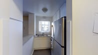 This single-floor kitchen is designed with a galley-style layout, maximizing efficiency within a compact footprint. The space is lined with white lower cabinetry and upper cabinets that extend toward the ceiling, offering ample storage on both sides of the narrow corridor. A stainless steel refrigerator anchors one end of the kitchen, complemented by a stainless steel dishwasher near the sink, giving the space a cohesive, modern appliance aesthetic. The countertops feature a speckled laminate surface, providing a neutral and functional work area. A double-basin sink is positioned beneath a window, which allows natural light to filter into the space, brightening the otherwise enclosed layout. The floors are finished with wood-look planking in a warm tone, adding a touch of warmth that contrasts with the crisp white walls and cabinetry. Flush-mount ceiling lighting ensures the space remains well-lit. The white walls and ceiling contribute to an open, clean feel, while the thoughtful use of vertical cabinet space ensures storage is prioritized throughout this efficiently arranged kitchen.