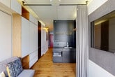 Located at 2221 S Clark St in Arlington, VA, this thoughtfully engineered micro-unit maximizes every inch of its compact footprint through clever, purpose-driven design. Warm hardwood flooring runs throughout, unifying the open layout and lending a welcoming ambiance to the space.

The combined living and dining area serves as the heart of the unit, anchored by a large window that floods the room with natural light and offers views of the surrounding neighborhood. A gray sofa, low coffee table, wall-mounted television, and built-in wooden credenza beneath the window make this area both functional and comfortable.

A curtained sleeping alcove provides a private retreat, housing a murphy-style bed framed by acoustic felt panels and an integrated wooden bookshelf — an efficient solution that preserves open floor space when not in use. Adjacent to the sleeping area, floor-to-ceiling modular cabinetry in white and natural wood tones offers generous storage.

The kitchenette is anchored by a dark pegboard feature wall fitted with hanging baskets, a stainless steel sink, and a built-in microwave, delivering a streamlined cooking space. A hallway connects the main living area to the entrance, closet, and bathroom.

The bathroom stands out with its playful design: penny-tile flooring, white square wall tiles, a navy blue vanity cabinet, and a bold mirror surround with orange-accented open shelving add a vibrant, modern character to the space.