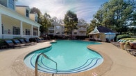 Located at 100 Preston Woods Trail in Sandy Springs, Georgia, this outdoor amenity area offers a well-appointed resort-style experience designed for relaxation and recreation. The centerpiece is a generously sized, freeform swimming pool with vivid turquoise water and decorative tiled lane accents along the pool floor, creating an inviting and visually striking focal point. Adjacent to the main pool is a separate hot tub, providing a dedicated space for unwinding. The expansive concrete sundeck surrounds the pool and is furnished with a row of cushioned lounge chairs, ideal for soaking up the Georgia sun. Outdoor seating areas with umbrella-shaded tables and chairs offer comfortable spots for socializing. A charming gazebo with a pitched roof anchors one end of the pool area, adding architectural interest and shade. The surrounding buildings feature a classic design combining brick accents, light-colored siding, and prominent white columns, contributing to the community's traditional aesthetic. Mature trees and manicured landscaping, including decorative shrubs and potted plants, frame the space and provide a natural, park-like setting. The overall ambiance is one of a thoughtfully designed, community-focused outdoor retreat that balances functionality with an attractive, welcoming atmosphere.