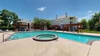 Located at 1773 Popps Ferry Rd in Biloxi, Mississippi, this outdoor amenity area is a well-appointed community pool complex designed for relaxation and recreation. The centerpiece is a generously sized rectangular swimming pool with crystal-clear blue water, complemented by an adjoining circular spa. The expansive concrete pool deck provides ample space for lounging, furnished with multiple sling-style chaise lounge chairs and patio seating arrangements ideal for socializing or unwinding under the Mississippi sun. Flanking the perimeter of the deck are several handsome wood-framed pergolas, adding architectural structure and shaded retreats along the walkways. A charming brick clubhouse building anchors one end of the space, featuring a hip roof, arched windows with green shutters, and elegant white columns supporting a covered porch. French doors open from the covered porch, suggesting an interior amenity or restroom facility. The surrounding landscape is lush with mature trees and manicured shrubs enclosed within a wrought iron fence, offering both privacy and a verdant backdrop. The overall setting conveys a resort-like ambiance perfectly suited to the warm Gulf Coast climate.
