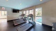 Located at 13831 Castle Blvd in Silver Spring, Maryland, this well-equipped fitness center offers a comprehensive workout environment on a single level. The space is thoughtfully organized into distinct cardio and strength training zones, providing a balanced facility for a full-body exercise routine.

The cardio area features multiple treadmills and an elliptical trainer, all positioned to take advantage of the large windows that line the walls. These windows offer views of the outdoor pool and lush surrounding trees, filling the space with natural light and creating an energizing atmosphere. A wall-mounted flat-screen television is also available to keep members entertained during cardio sessions.

The strength training zone is equally well-appointed, housing a variety of cable and weight machines, a lat pulldown station, a bench press, and additional cable resistance equipment. Structural columns define the layout of this area, while multiple windows continue to bring in natural light. The flooring throughout is durable rubber matting, ideal for a fitness environment.

Recessed lighting in the smooth ceiling supplements the abundant natural light, ensuring the space is well-lit at all times. A glass-paneled door provides access to and from the facility. The overall ambiance is clean, bright, and functional, making this fitness center a practical and inviting amenity.