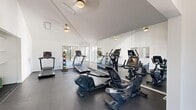 Located at 632 E James Pointe Dr in Murray, Utah, this single-room fitness center offers a well-equipped workout space designed for efficient, functional exercise. The room features vaulted ceilings that lend a sense of openness, complemented by crisp white walls and durable rubber flooring suited for high-traffic athletic use. Natural light pours in through multiple windows along one wall, providing views of surrounding greenery and creating a bright, energizing atmosphere. A large wall-mounted mirror spans one side of the room, visually expanding the space and aiding in form correction during workouts. The cardio section is well-stocked with multiple treadmills, elliptical trainers, and a stationary bike, catering to a variety of aerobic preferences. A Hoist functional trainer cable machine serves as the centerpiece for strength training, accompanied by a medicine ball rack offering a range of weighted options for functional fitness. A wall-mounted television provides entertainment during workouts, while a stability ball and additional accessories round out the available equipment. The space is accessed through a glass-paneled entry door and maintains a clean, minimalist aesthetic throughout, making it a practical and inviting facility for residents seeking convenient on-site fitness amenities.