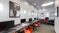 This contemporary business center is designed to provide a productive and inspiring work environment. The space features a long, dark granite-topped workstation surface running along the wall, accommodating multiple iMac desktop computers, each paired with a keyboard and mouse. Ergonomic orange rolling chairs add a vibrant pop of color against the neutral gray walls and patterned carpet, creating an energetic yet professional atmosphere. The room is well-lit with a combination of recessed ceiling lights and stylish white pendant lights that hang at varying heights, contributing to a modern aesthetic. The ceilings are notably tall, enhancing the open and airy feel of the space. The walls are thoughtfully adorned with large black-and-white framed portraits and motivational quote prints — including recognizable figures such as Albert Einstein and references to iconic speeches — reinforcing an atmosphere of innovation and inspiration. A secondary workstation area is visible toward the back of the room, offering additional workspace. The overall design balances functionality with a polished, contemporary style, making it an ideal setting for focused work or collaborative productivity.