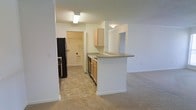 This single-floor residential space offers a practical and comfortable open-concept layout. The kitchen is centrally positioned and well-equipped, featuring light wood cabinetry, a black refrigerator, a dishwasher, and a countertop that extends into a peninsula-style breakfast bar — ideal for casual dining or additional prep space. The tile flooring in the kitchen provides a clean, easy-to-maintain surface, while the adjoining living area transitions seamlessly into plush wall-to-wall carpeting, adding warmth underfoot. The open flow between the kitchen and living space creates a sense of connectivity and ease of movement throughout. Neutral tones across the walls, cabinetry, and countertops give the space a bright, cohesive feel. A dedicated laundry or utility area is visible toward the rear of the kitchen, offering convenient in-unit functionality. Doorways leading to additional rooms suggest a thoughtful layout that balances shared and private spaces. Overhead lighting is present throughout, ensuring consistent illumination. The overall design is clean and straightforward, making it a highly functional living environment suited to everyday comfort.