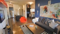 This single-floor space showcases a vibrant, contemporary design with an open-concept layout that connects the living, dining, and kitchen areas into one cohesive environment. The living area is anchored by a bold navy blue accent wall, which provides a dramatic backdrop for a large abstract painting and a comfortable gray sofa adorned with decorative pillows. A striking orange accent chair adds a pop of color, complementing the chevron-patterned area rug and a sleek rectangular coffee table beneath.

Hardwood flooring runs throughout, lending warmth and continuity across the entire space. The kitchen features dark cabinetry with a kitchen island, pendant lighting overhead, and an adjoining dining area with a compact table and modern chairs. A large arc floor lamp elegantly bridges the living and dining zones, providing ambient lighting.

The open layout maximizes the sense of space, while the curated mix of bold colors, modern furnishings, and layered lighting creates an energetic yet inviting atmosphere throughout. The overall design reflects a well-thought-out use of space with a distinctly urban, contemporary aesthetic.