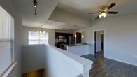 This single-floor space showcases a thoughtfully designed open-concept layout that seamlessly connects the kitchen, dining, and living areas. The kitchen is a standout feature, outfitted with dark cabinetry, stainless steel appliances including a refrigerator, stove, and microwave, and a sleek kitchen island with a sink and light countertops that provide generous prep and casual dining space. Track lighting overhead illuminates the kitchen, while a pendant chandelier adds a warm, decorative touch above the dining area. The vaulted ceiling creates an impressive sense of vertical openness and airiness throughout the main living space, enhanced further by a ceiling fan with a light fixture that promotes both comfort and circulation. Rich, dark hardwood-style flooring runs throughout, grounding the space with warmth and contrast against the light-colored walls and countertops. A large window in the dining area welcomes abundant natural light. A hallway leads to additional rooms, including what appears to be a bathroom and closet or bedroom spaces, maintaining a functional and well-organized floor plan. Overall, the space conveys a clean, contemporary aesthetic with modern finishes and a bright, welcoming ambiance.