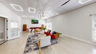 Located at 1800 Boydton Plank Rd in Petersburg, Virginia, this single-floor multipurpose space offers a well-appointed and inviting environment designed for both leisure and productivity. The open-plan layout is finished with light wood-style flooring and crisp white walls, creating a clean, bright atmosphere throughout.

The space is divided into distinct functional zones. A comfortable lounge area features a bold red sofa accented with colorful throw pillows, paired with stylish white and gold accent chairs arranged around a round ottoman coffee table atop a decorative blue and grey area rug. A wall-mounted flat-screen television adds an entertainment focal point. Pendant lighting and geometric LED ceiling fixtures illuminate the space with a modern, layered lighting design.

A dedicated recreation zone houses a full-size billiards table with a red felt surface, complemented by bar-height stools — ideal for casual social gatherings. Large windows with horizontal blinds bring in generous natural light and offer views of the surrounding greenery.

A workspace area rounds out the room, featuring a sleek industrial-style desk with a desktop computer, providing a functional setting for focused tasks. Abstract wall art adds a creative touch. The overall ambiance is energetic yet welcoming, making this space well-suited for community engagement, recreation, and collaborative work.