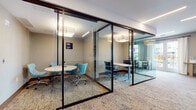 Located at 40 Bennett in Englewood, NJ, this single-floor workspace offers a thoughtfully designed environment tailored for collaboration and focused work. The layout features multiple glass-walled work rooms framed in sleek dark metal, providing visual transparency while maintaining acoustic separation. These enclosed rooms are furnished with round and rectangular tables paired with contemporary seating in teal and blue tones, making them well-suited for small group meetings or private work sessions.

An open meeting area accommodates larger gatherings, featuring long wooden conference tables surrounded by teal velvet chairs with gold-accented legs, complemented by a vibrant abstract wall artwork that adds a creative focal point. Recessed linear lighting and patterned carpeting throughout contribute to a polished, professional atmosphere.

Additional workstations with glass partitions offer semi-private individual workspace, while a shared utility area near large windows and French doors provides access to printing and administrative amenities. Natural light flows in through generous windows and glass doors, offering views of the surrounding outdoor courtyard. The overall design balances openness with privacy, creating a versatile and modern professional environment.
