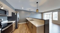 This single-floor space showcases a well-appointed kitchen designed with a clean, modern aesthetic. Light wood cabinetry lines the walls and base units, providing ample storage while contributing to a warm, contemporary feel. The countertops feature a speckled stone surface in neutral tones, offering both durability and visual appeal. A stainless steel sink with a modern arc-neck faucet is integrated seamlessly into the counter run, complemented by a stainless steel refrigerator with a built-in water dispenser nearby. Two distinctive pendant lights hang from the ceiling — each with a metallic, banded cylindrical shade — adding character and focused task lighting over the counter space. Recessed lighting supplements the ambient illumination throughout. The flooring consists of warm-toned hardwood planks that run continuously, enhancing the sense of openness. Cool grey walls provide a neutral backdrop that contrasts tastefully with the wood tones. A window at the far end of the kitchen allows natural light to filter into the space, brightening the overall atmosphere. The layout is efficient and functional, with a corridor-style design that maximizes counter and storage space.