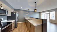 This single-floor space showcases a well-appointed kitchen designed with a clean, modern aesthetic. Light wood cabinetry lines the walls and base units, providing ample storage while contributing to a warm, contemporary feel. The countertops feature a speckled stone surface in neutral tones, offering both durability and visual appeal. A stainless steel sink with a modern arc-neck faucet is integrated seamlessly into the counter run, complemented by a stainless steel refrigerator with a built-in water dispenser nearby. Two distinctive pendant lights hang from the ceiling — each with a metallic, banded cylindrical shade — adding character and focused task lighting over the counter space. Recessed lighting supplements the ambient illumination throughout. The flooring consists of warm-toned hardwood planks that run continuously, enhancing the sense of openness. Cool grey walls provide a neutral backdrop that contrasts tastefully with the wood tones. A window at the far end of the kitchen allows natural light to filter into the space, brightening the overall atmosphere. The layout is efficient and functional, with a corridor-style design that maximizes counter and storage space.