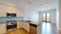 This well-appointed single-floor residence offers a thoughtfully designed open-concept layout that seamlessly connects the kitchen and living areas. The kitchen is a standout feature, equipped with a full suite of stainless steel appliances including a gas range, oven, built-in microwave, French door refrigerator with ice maker, and dishwasher. White flat-panel cabinetry reaches toward the ceiling, providing generous storage, while a light blue subway tile backsplash adds a subtle pop of color. A central island with granite countertops and an undermount sink serves as both a functional workspace and a natural focal point, illuminated by elegant pendant lighting. Track lighting runs throughout the main living area, which boasts warm hardwood flooring and large French doors that flood the space with natural light and open to views of the surrounding neighborhood. The bedroom features plush neutral carpeting and a stylish panel door leading to a dedicated closet. The bathroom is finished with matching granite countertops, white cabinetry, and a glass-enclosed shower. A stacked in-unit washer and dryer adds everyday convenience. The overall aesthetic is clean, contemporary, and bright, creating a cohesive and comfortable living environment.