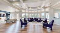 Located at 3240 SW 34th St in Ocala, Florida, this single-floor clubhouse facility is thoughtfully designed to serve as a sophisticated community gathering space. The centerpiece of the building is a dramatic rotunda lounge, defined by soaring ceilings, clerestory windows wrapping the curved walls, and an abundance of natural light that highlights the warm hardwood-style flooring below. Deep navy-blue upholstered seating, a glass-top coffee table, and a patterned area rug create an inviting and polished atmosphere, with direct views of an outdoor pool and covered patio beyond the expansive windows.

Adjacent to the lounge is a well-appointed business center and media room, featuring a glass-top workstation, built-in shelving set against a bold blue accent wall, a wall-mounted flat-screen television, and comfortable seating — all conducive to both productivity and relaxation. The facility also includes a fully equipped kitchen with white cabinetry, granite countertops, stainless steel appliances, and a window overlooking the pool area.

A separate dining and social area showcases coffered ceilings with an elegant chandelier, double French doors opening to the outdoors, and stylish high-back chairs paired with bistro-style tables. Throughout the space, consistent design elements — warm flooring, refined furnishings, and abundant natural light — create a cohesive, upscale ambiance.