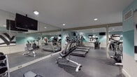 The Penniman Park fitness center is a well-equipped, single-level exercise facility designed to support a comprehensive range of workout routines. The room is finished with durable rubber flooring in a speckled grey tone, providing a stable and resilient surface suited for high-traffic use. Large floor-to-ceiling mirrors line multiple walls, visually expanding the space and allowing users to monitor their form during exercise.

The cardio section features multiple Cybex treadmills positioned in rows, offering ample capacity for simultaneous use. A recumbent stationary bike provides a low-impact cardiovascular option, while a multi-station cable and weight machine supports a variety of strength-training exercises. Free weights and an adjustable bench are available for targeted resistance training, and a freestanding Century punching bag adds a functional combat fitness element.

The walls are painted in a two-tone scheme combining soft teal and neutral white, contributing to a clean, energetic atmosphere. Recessed ceiling lighting ensures consistent illumination throughout the room, and wall-mounted flat-screen televisions are positioned for visibility from multiple workout stations. Decorative wall art adds a polished, finished aesthetic to the overall environment.