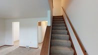 This three-floor home offers a practical and straightforward layout designed for comfortable everyday living. The main level features a bright living area with large windows dressed in horizontal blinds, allowing natural light to fill the carpeted space. The kitchen is well-equipped with warm wood cabinetry, dark granite-style countertops, tile flooring, and white appliances including a dishwasher, electric range, and over-the-range microwave, complemented by a window above the sink. A compact bathroom on one of the upper levels includes a pedestal sink, a recessed medicine cabinet with vanity lighting, and a towel bar, finished with textured walls. The upper floors host multiple carpeted bedrooms, each outfitted with ceiling fans and windows offering views of surrounding greenery, providing a calm and airy atmosphere. A carpeted staircase with a warm wood railing and banister connects the levels, accented by vaulted ceiling details that add vertical dimension to the stairwell. The lower level presents an unfinished basement with exposed wooden joists, concrete block walls, and a utility sink, offering generous potential for storage, a workshop, or future finishing. Overall, this home provides a clean, functional foundation with room to personalize.