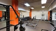 This single-floor fitness facility is designed to support a full range of strength training activities. The space features bold orange accent walls contrasted against neutral tones, creating an energetic atmosphere suited to an active environment. Rubber flooring throughout provides durability and safety underfoot, while flush-mounted fluorescent panel lighting ensures the area is well-lit for workouts.

A substantial dumbbell rack lines one wall, stocked with a wide selection of free weights to accommodate various training levels. Multiple adjustable weight benches are positioned throughout the floor, offering versatility for different exercises. A wall-mounted mirror along one side enhances the sense of space while allowing users to monitor their form. Additional strength equipment, including a cable or multi-function station, is positioned toward the rear of the room.

Exposed ceiling infrastructure adds an industrial character to the space. The layout is open and functional, with equipment arranged to maximize usable floor area and allow comfortable movement between stations. Overall, the space presents a practical, no-frills training environment focused on delivering effective strength and conditioning capabilities.