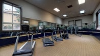 This single-level fitness center is designed to support a comprehensive workout experience in a bright, open environment. The space is outfitted with a variety of cardio equipment, including multiple True-brand treadmills and elliptical machines, as well as strength training cable machines and free weight benches, providing users with a full range of exercise options.

Large windows line the walls, inviting generous amounts of natural light and offering views of the surrounding greenery, creating an airy and motivating atmosphere. Full-length mirrors with navy blue base panels span multiple walls, visually expanding the space and allowing users to monitor their form during workouts. Wall-mounted televisions add to the convenience and entertainment options available.

The flooring throughout consists of durable, textured carpet tiles well-suited for a high-traffic fitness environment. Ceiling fans are installed to maintain comfortable airflow, complementing the recessed and panel lighting overhead. The neutral-toned walls and drop ceiling contribute to a clean, functional aesthetic. The open layout ensures ample room for movement between equipment, making this fitness center both practical and inviting for daily use.