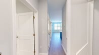 This well-appointed unit in Swanzey, NH offers a thoughtfully designed single-floor layout with one bedroom, a versatile den, and one full bathroom. The open-concept living and kitchen area creates a welcoming, airy atmosphere anchored by light wood-look flooring and soft blue-gray walls that carry consistently throughout the home. The modern kitchen is a standout feature, showcasing crisp white cabinetry, a subway tile backsplash, quartz countertops, a central island with pendant lighting, and a full suite of stainless steel appliances including a range, over-the-range microwave, dishwasher, and refrigerator. The living room is bright and open, with large windows that invite natural light and offer views of the surrounding neighborhood. The primary bedroom is generously sized, featuring a wide window with outdoor views and ample room for furnishings. The den provides flexible additional space ideal for a home office or guest room, also benefiting from natural light. The full bathroom features marble-look tile flooring, a soaking tub with shower, and clean, contemporary finishes. A dedicated closet adds practical storage. The wide, well-lit hallway connects all rooms seamlessly, reinforcing the unit's clean and modern aesthetic.