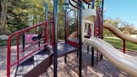 This outdoor playground is a well-equipped recreational space designed to offer a variety of activities for children. The centerpiece is a multi-level play structure constructed with durable dark metal posts and bold red railings, providing a visually striking and sturdy framework. The structure includes a winding slide, a spiral enclosed slide, and multiple elevated platforms connected by metal grate staircases and ramps, promoting both accessibility and active play.

Along the perimeter of the main structure, interactive play panels are mounted, offering sensory and educational engagement. An overhead horizontal ladder bar extends from the main unit, adding an additional physical challenge for older children. A standalone red bench nearby provides a comfortable resting spot for supervising adults.

The ground throughout the play area is covered in wood chip surfacing, offering a cushioned, safety-conscious base beneath the equipment. The playground is bordered by lush green grass and framed by a dense backdrop of mature deciduous trees, creating a natural and inviting atmosphere. Views from the playground reveal surrounding residential buildings and open green lawns, situating this space within an active community setting. Ample natural light floods the area, making it a bright and welcoming environment throughout the day.