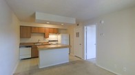 This single-story space features an open-concept design that seamlessly connects the kitchen and living area. The kitchen is equipped with warm honey-toned wood cabinetry, a white electric range, a white refrigerator, and a built-in dishwasher. A peninsula with a laminate countertop extends into the living space, providing additional prep area and serving as a natural divider between the two zones. Recessed lighting beneath a soffit above the kitchen adds focused illumination to the workspace, complemented by an under-cabinet light above the range. The sink is positioned along the base cabinetry facing the living area, enhancing the open feel. Light-toned carpeting covers the living area, while the kitchen features easy-to-clean flooring. The walls are finished in a clean, neutral beige palette throughout. A textured ceiling adds subtle visual interest overhead. Two doors visible from the main space provide access to additional rooms or storage, and a louvered-panel door suggests a utility or closet area. The overall layout is practical and well-organized, making efficient use of the available space.